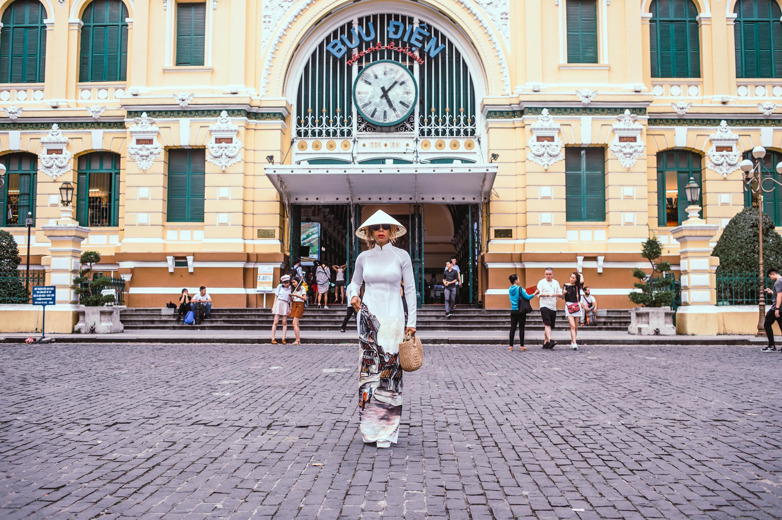 A woman in traditional Vietnamese Ao Dai stands outside Ho Chi Minh City's historic Central Post Office.